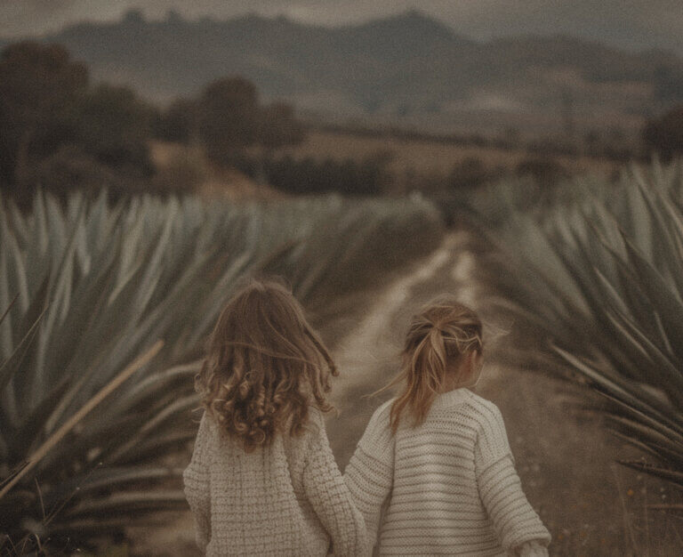 A photo of two little girls, one with long brown hair and the other with short blonde hair, in loose white sweaters over red pants running on an old dirt road surrounded by agave plants.